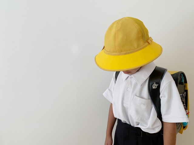 alt="帽子をかぶりランドセルを背負って下を向く、小学校低学年の子ども。学童に行きたくなくて気持ちが沈んでいる様子"